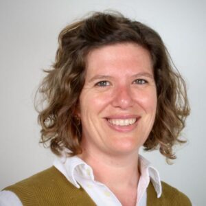 Smiling woman with shoulder-length curly light brown hair, wearing a white shirt and mustard cardigan, against a neutral background.