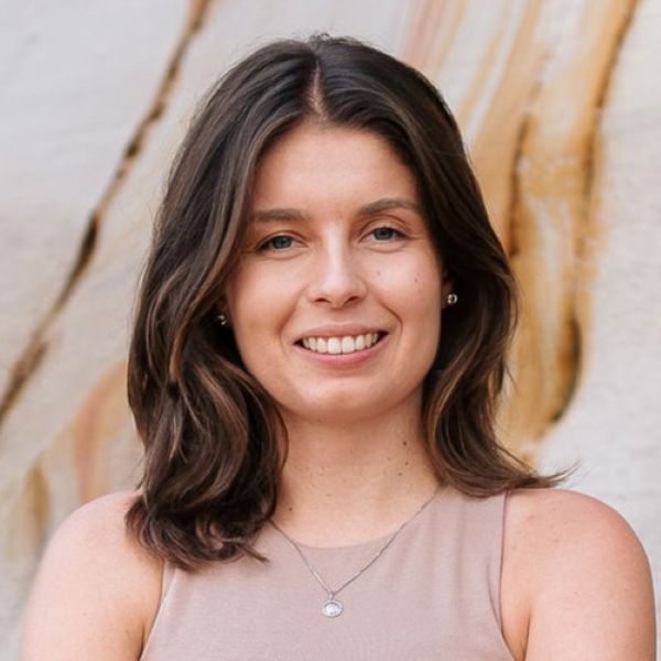 Portrait of a smiling woman with shoulder-length brown hair wearing a beige sleeveless top, standing in front of a light stone wall.