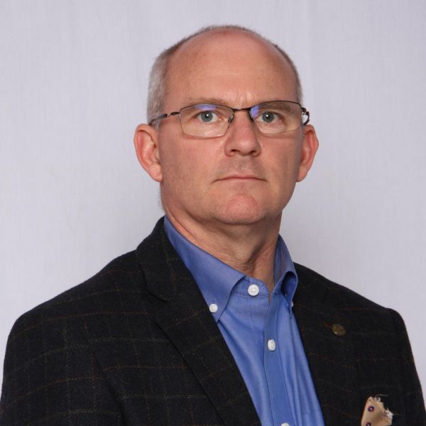 Head-and-shoulders portrait of a middle-aged man with glasses, wearing a blue button-down shirt and dark blazer.
